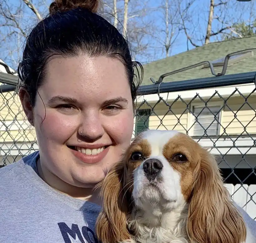A smiling team member holding a Cavalier King Charles spaniel outside the kennel building